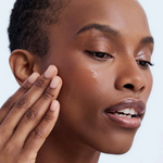 Woman applying cream to her face with a jar of cream in her hand against a light background