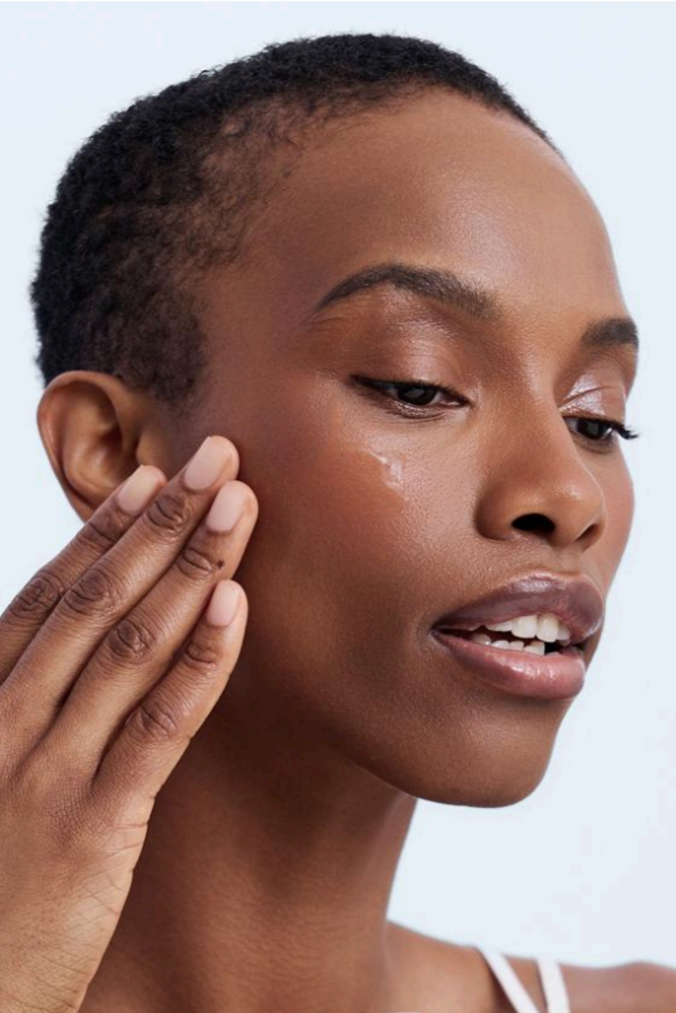 Woman applying cream to her face with a jar of cream in her hand against a light background